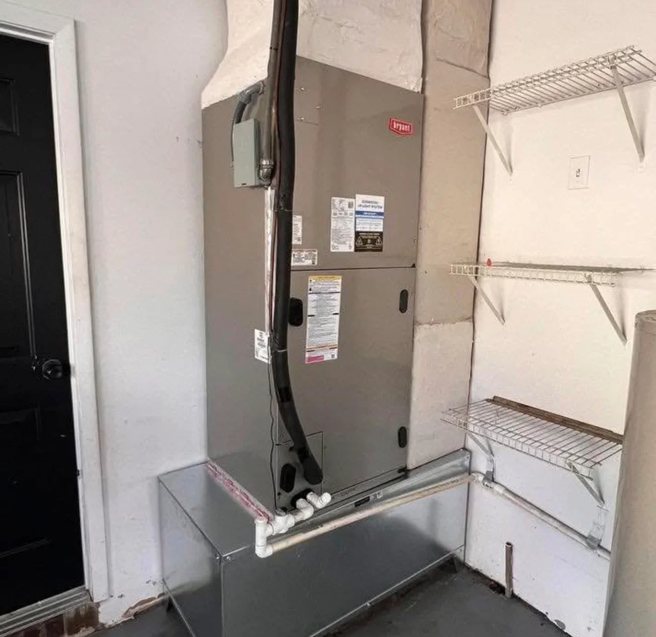 Installed furnace with metal platform base, black ductwork, and wire shelving in mechanical room